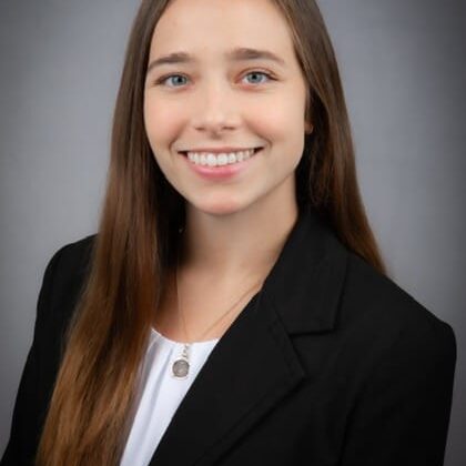 Professional headshot of a smiling young woman with long brown hair, wearing a black blazer and white blouse.